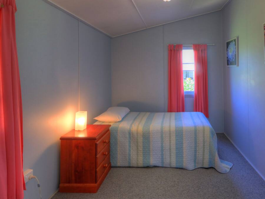 Un dormitorio con cama, mesita de noche, ventana con cortinas e imagen colgada en la pared.