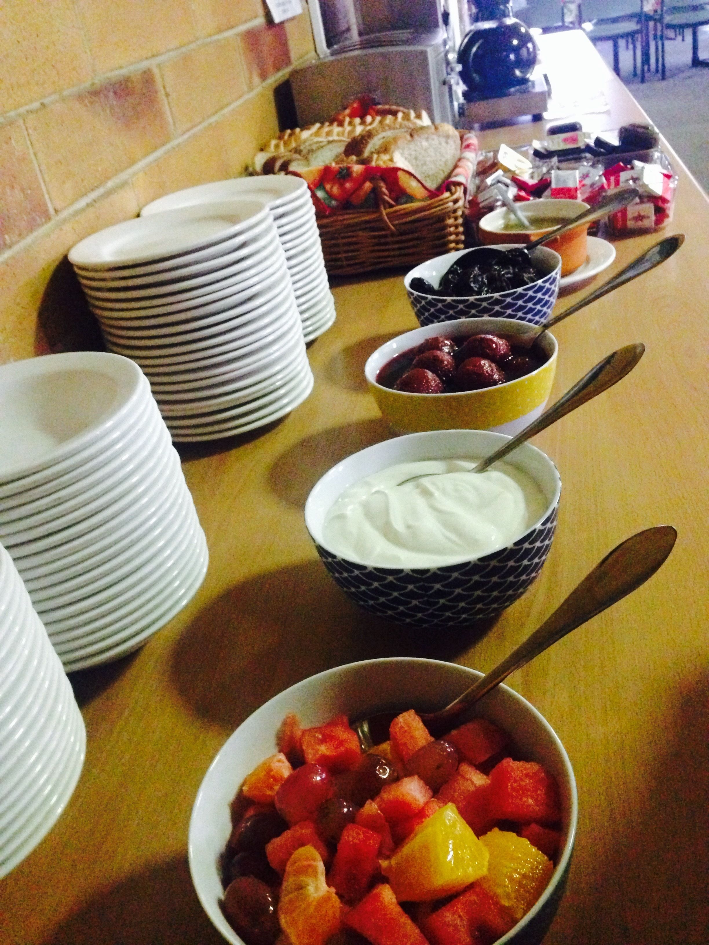 Una mesa de buffet con diversos platos, incluyendo cuencos de fruta, yogur y otros alimentos, además de platos apilados y utensilios de servir.