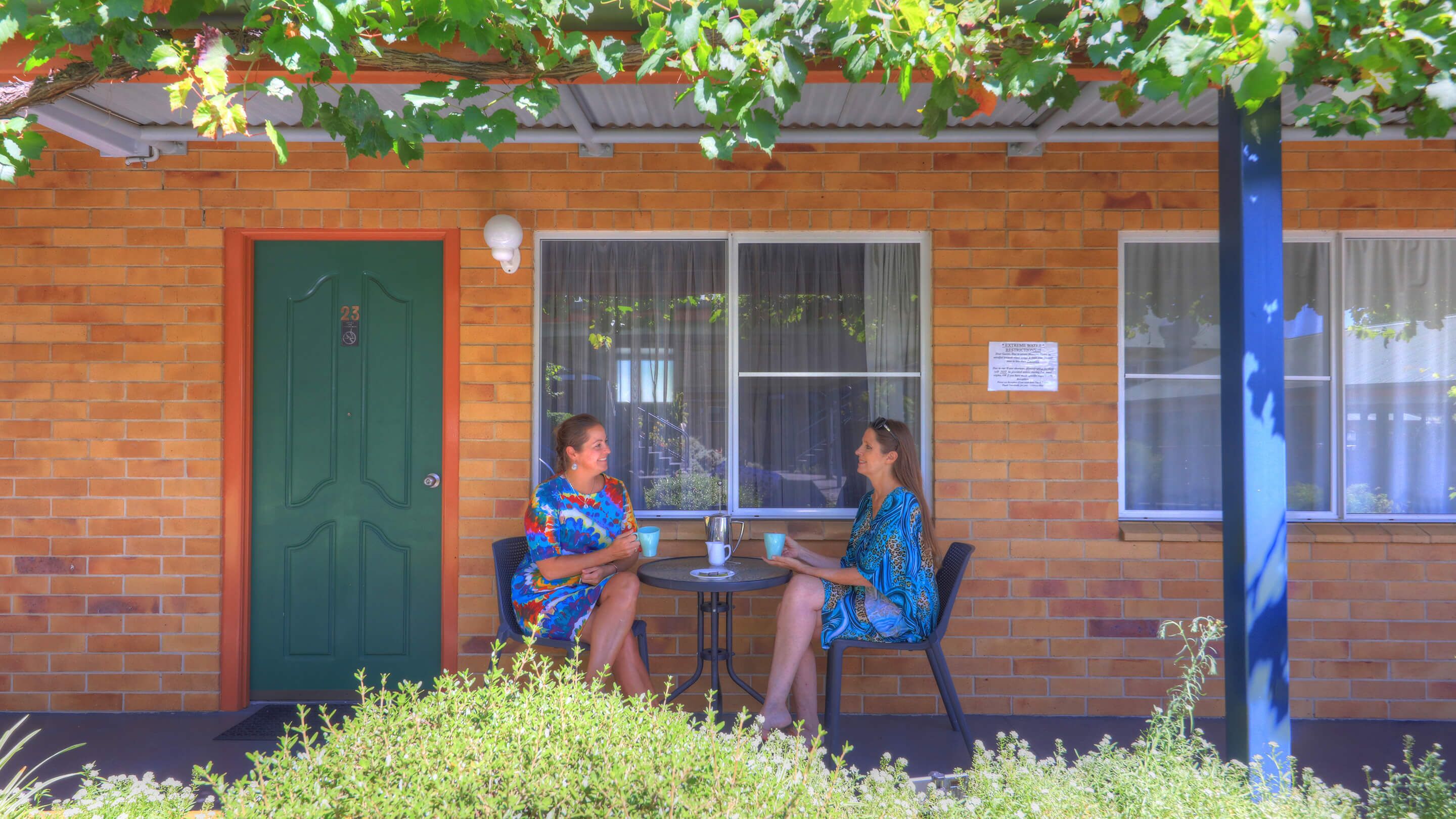 Two people seated at a small round table outside a brick building with a green door and windows.