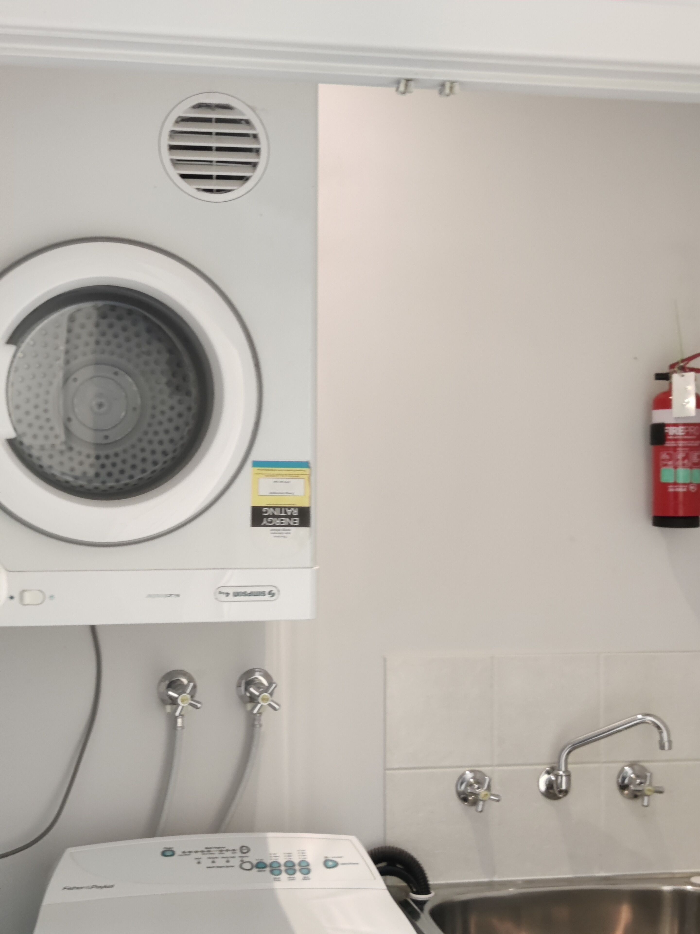 A compact laundry area with a front-loading washing machine, a wall-mounted fire extinguisher, and a sink with a faucet.