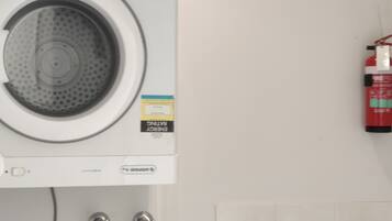 A compact laundry area with a front-loading washing machine, a wall-mounted fire extinguisher, and a sink with a faucet.