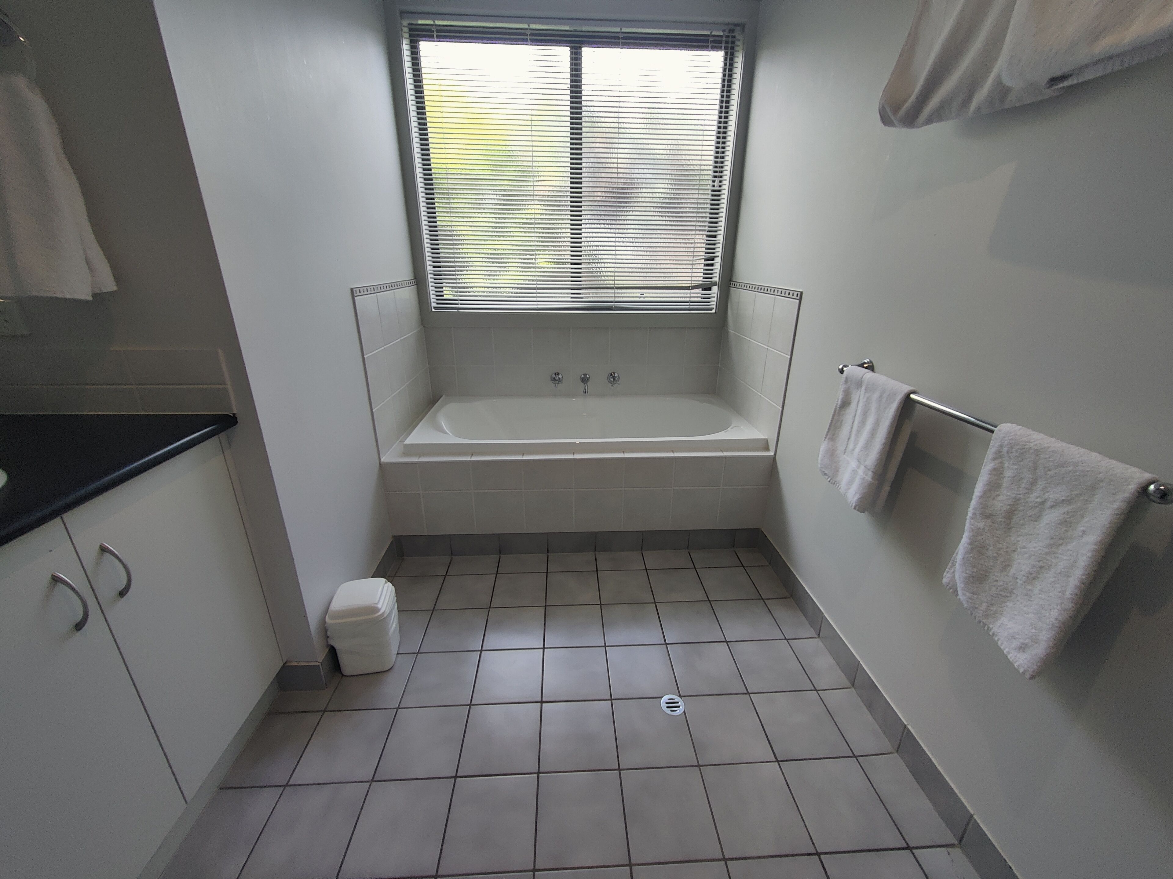 A small, clean bathroom with a bathtub, a window with blinds, white tiles, and a towel rack.