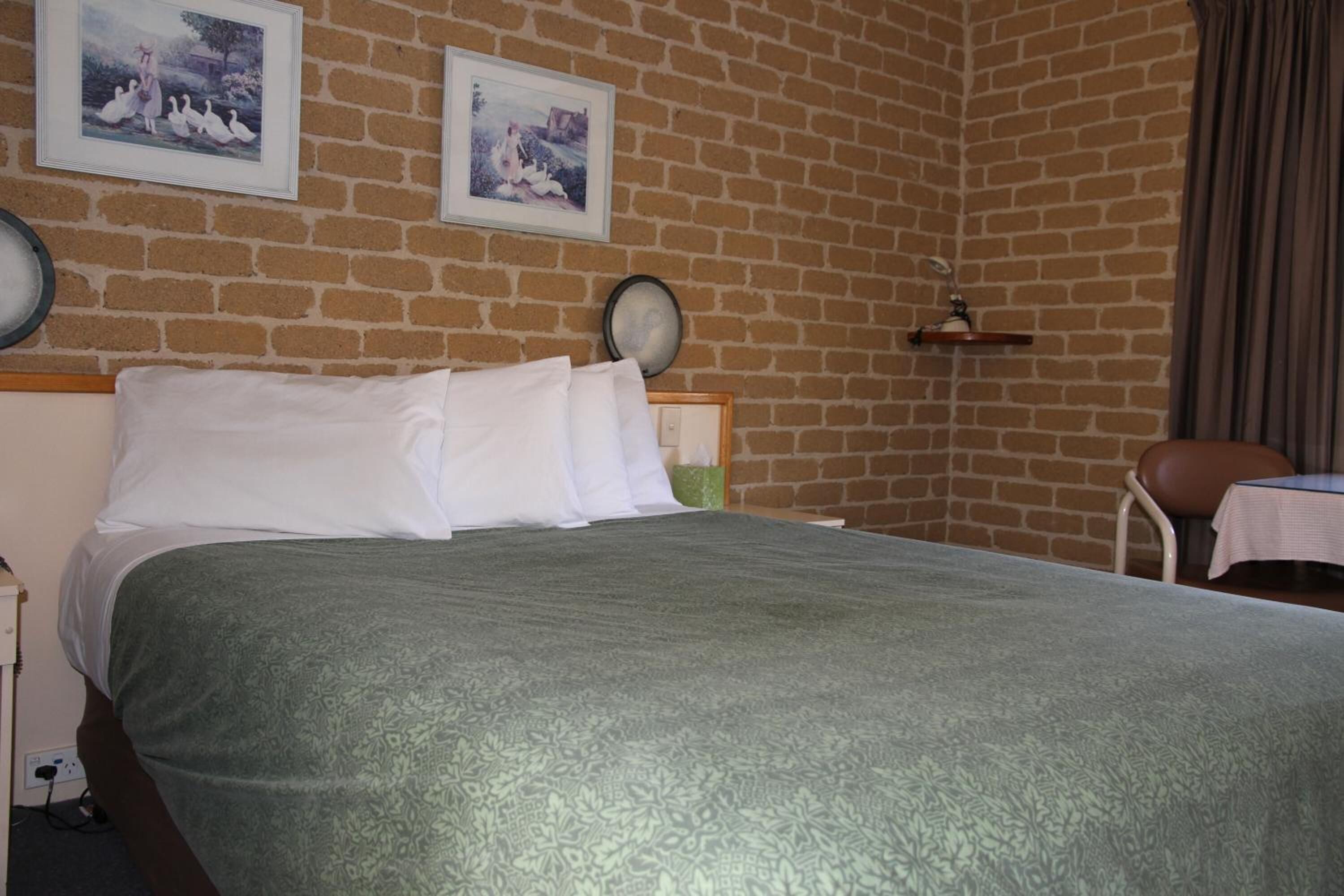 A hotel room with a brick wall, a bed with white linens, a green patterned bedspread, and two framed pictures on the wall.