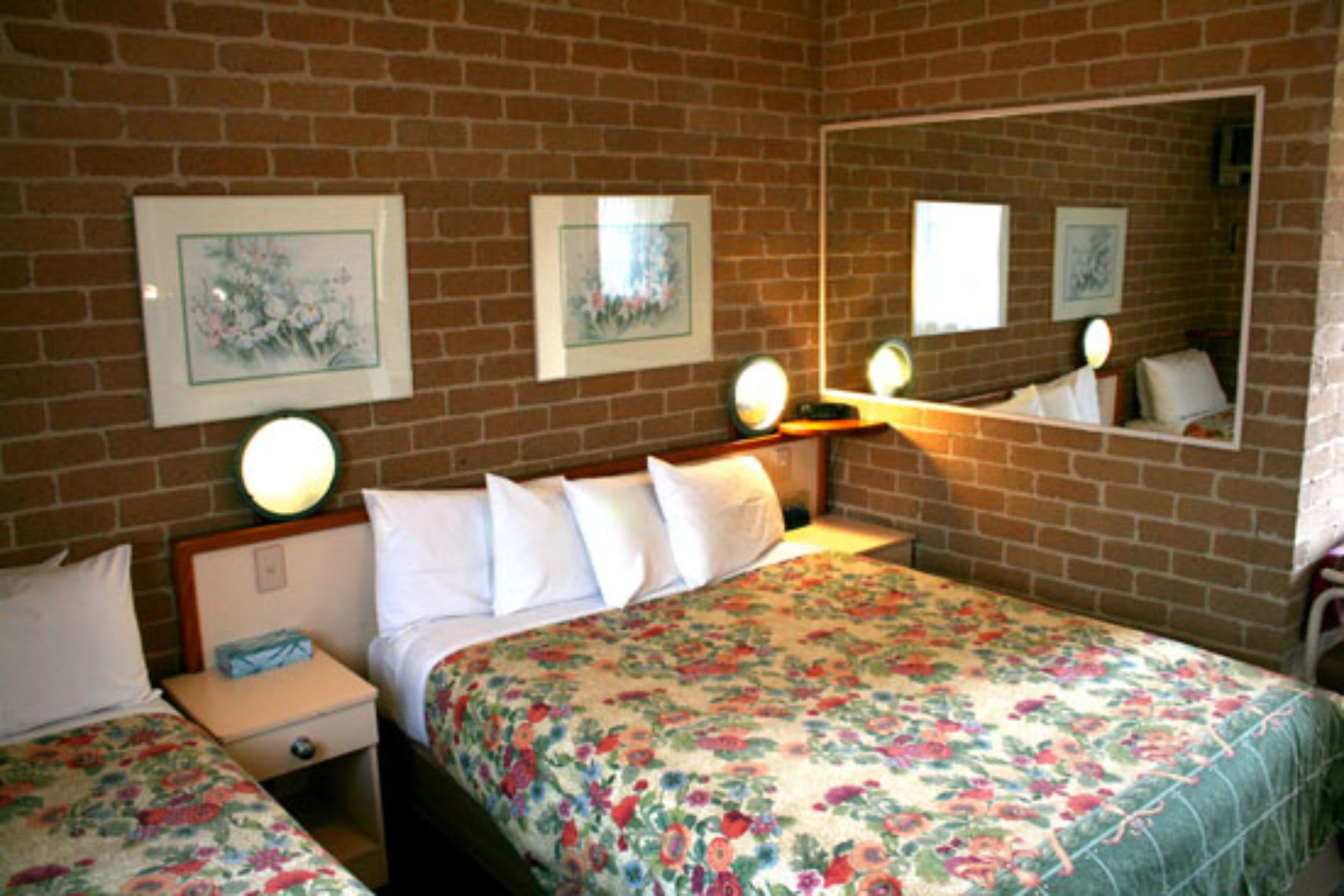 Una habitación de hotel con pared de ladrillo, dos camas con ropa de cama floral, mesitas de noche con lámparas y cuadros enmarcados en la pared.