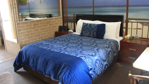 A bedroom with a bed, a nightstand, a mirror, and a view of the ocean. - Grand Manor Motor Inn - Queanbeyan (Queanbeyan East)