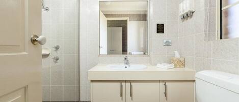A modern bathroom with a white sink, a mirror above it, and a toilet. There is a shower area to the left and a door leading to another room.