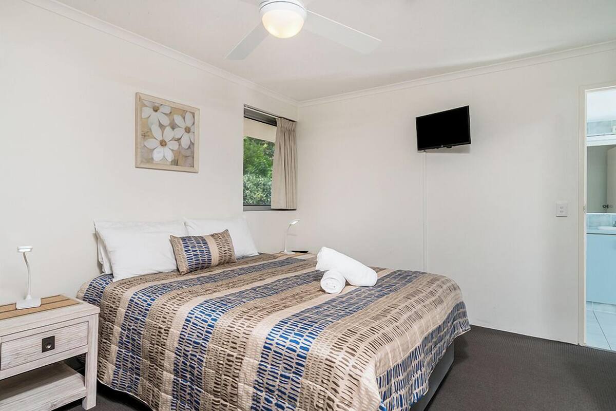 A bedroom with a bed, a nightstand, a TV, and a window.