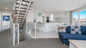 A modern kitchen with white cabinets, a dining area with white chairs, and a blue sofa with a patterned pillow. A wooden staircase leads to an upper level. - Rainbow Bay Resort (Coolangatta)