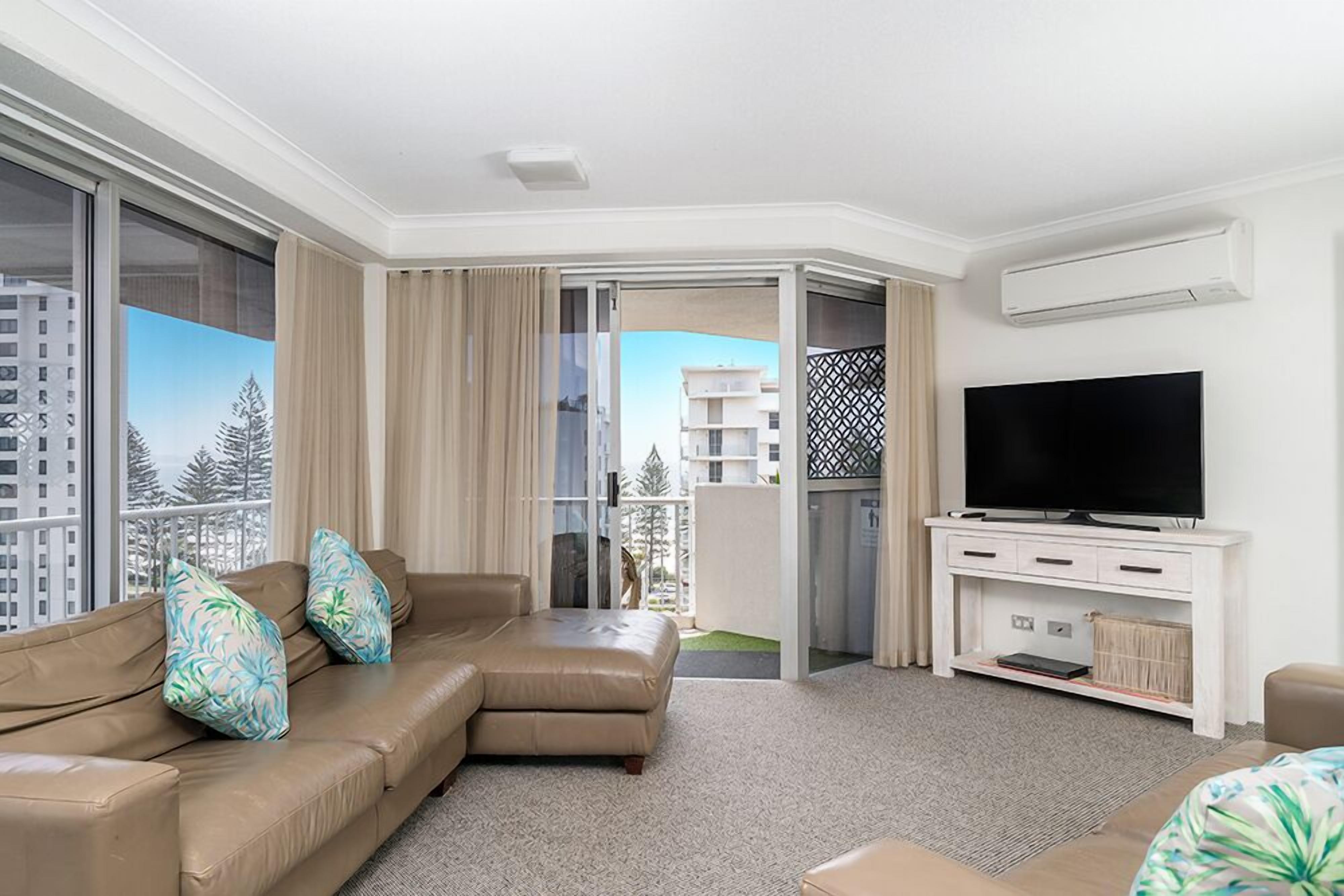 A modern hotel room with a beige sofa, a flat-screen TV, and a balcony with a view of buildings and trees.