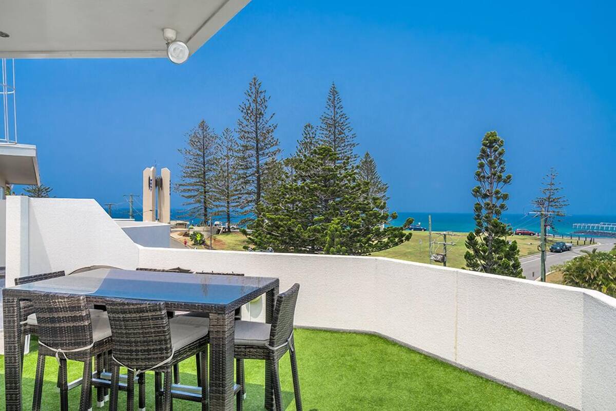A balcony with a table and chairs, overlooking a coastal area with trees and a clear blue sky.