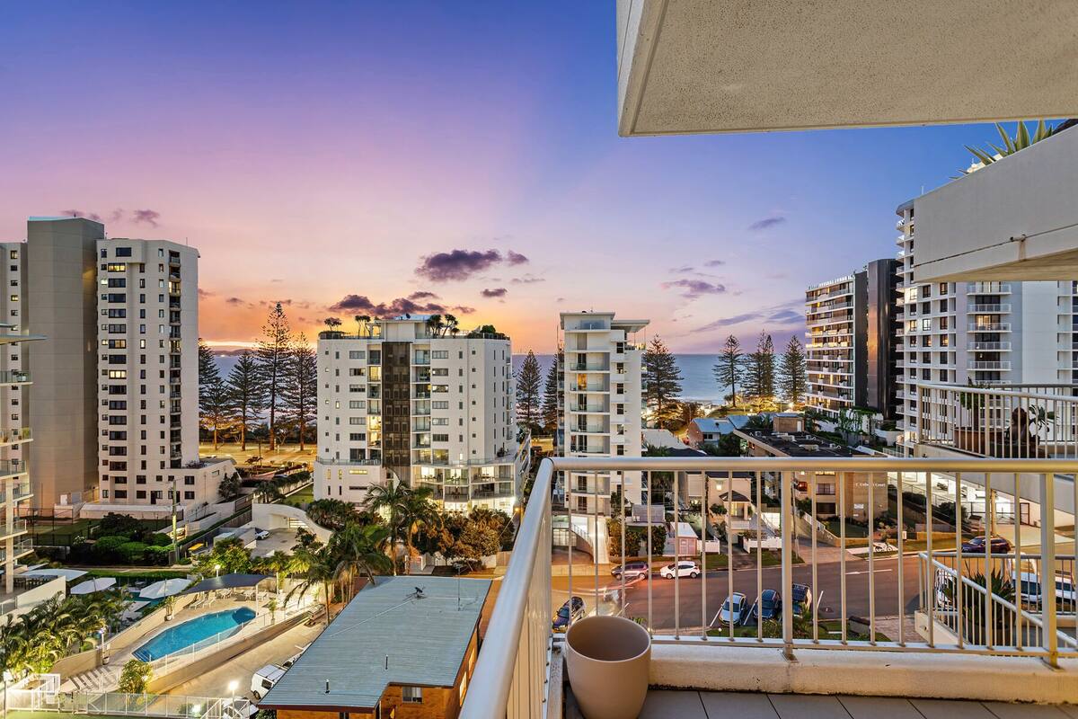 A high-rise building with multiple balconies, a swimming pool, and a view of the ocean at sunset.