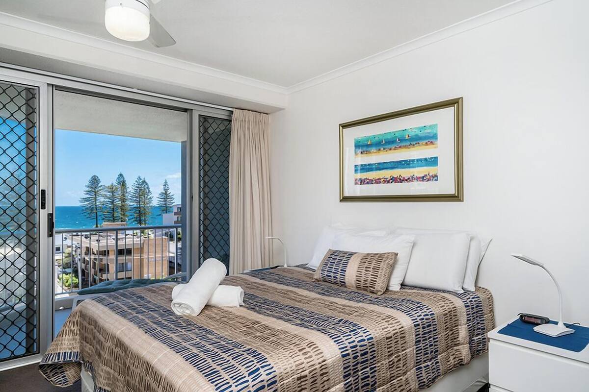 A hotel room with a bed, a nightstand, a framed picture, and a balcony with a view of the ocean and trees.