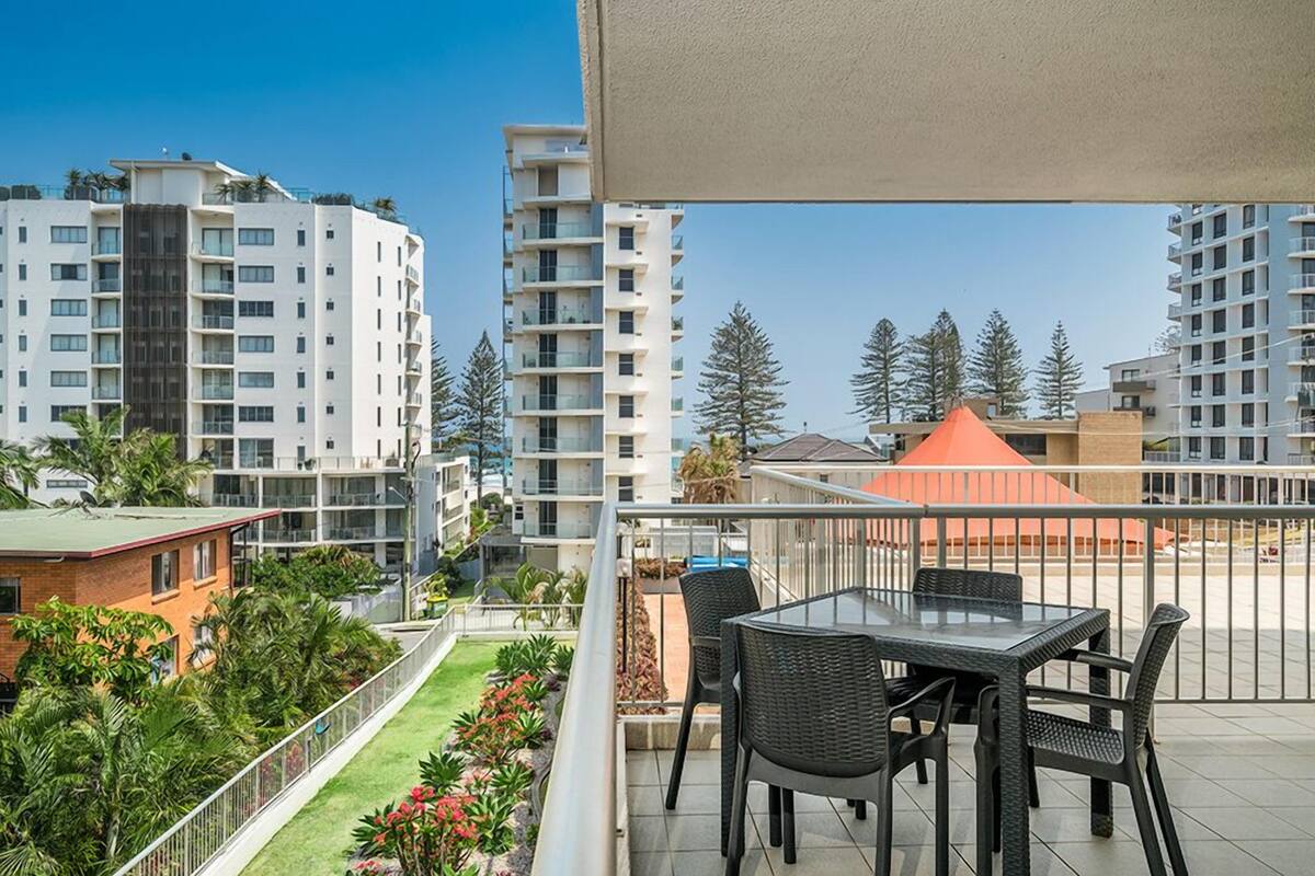A balcony with a table and chairs, overlooking a garden and buildings.