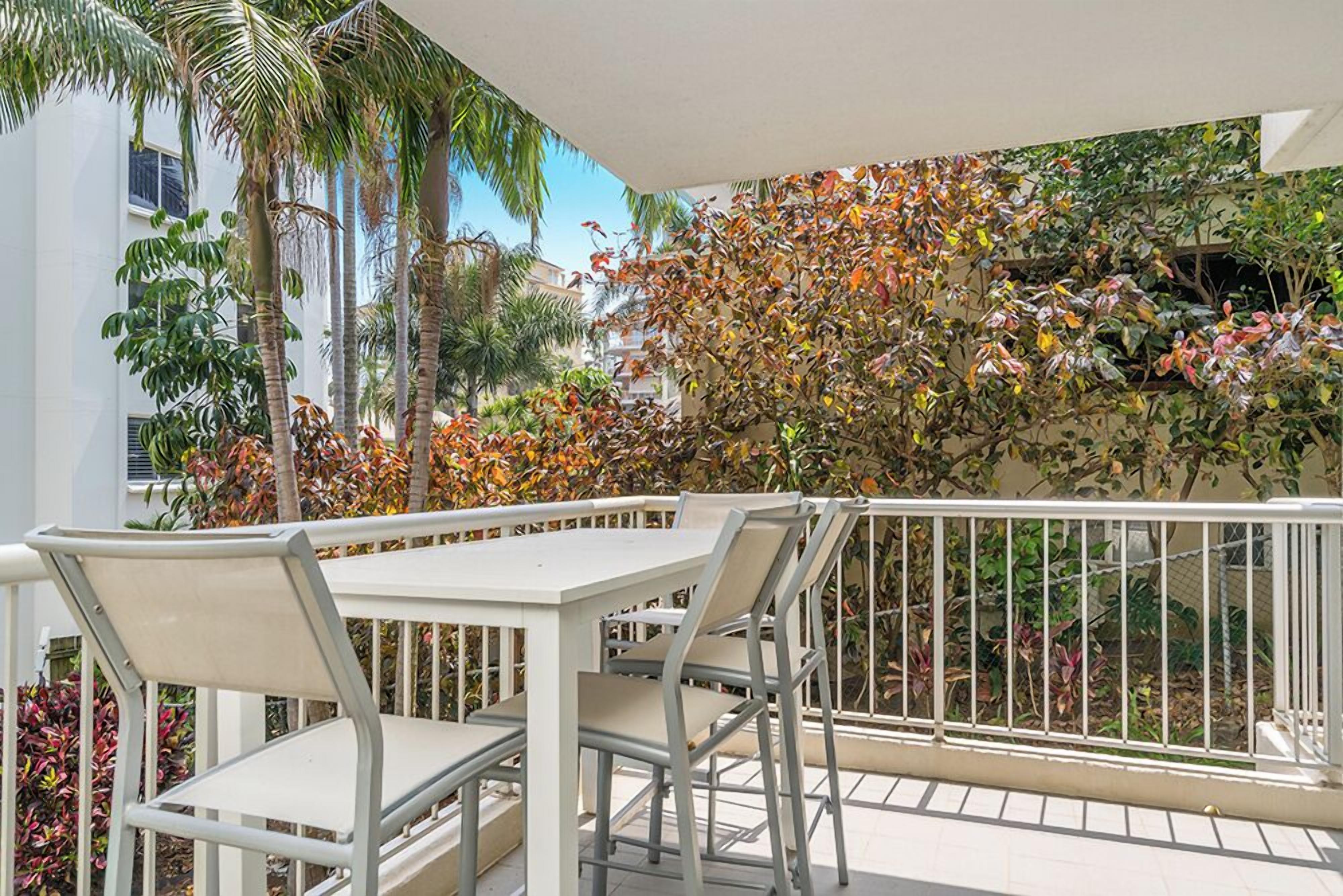 Ein Balkon mit weißem Tisch und Stühlen, umgeben von einer weißen Brüstung, mit Blick auf einen Garten mit verschiedenen Pflanzen und Bäumen.