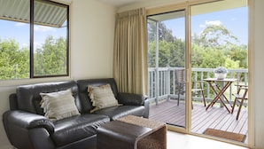 A living room with a black leather sofa, two cushions, and a woven ottoman. There is a sliding glass door leading to an outdoor deck with a table and chairs. - Mystery Bay Cottages (Mystery Bay)