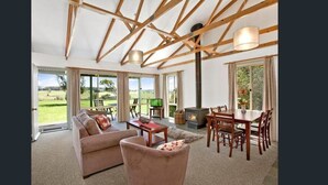A spacious living room with a sofa, coffee table, and dining area. A large window offers a view of the outdoors. - Mystery Bay Cottages (Mystery Bay)