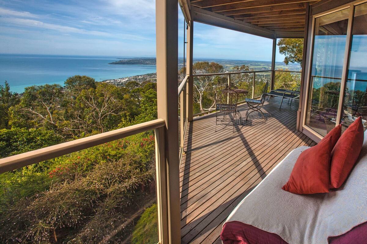 A balcony with a wooden deck, a sofa with red cushions, and a view of the sea and landscape.