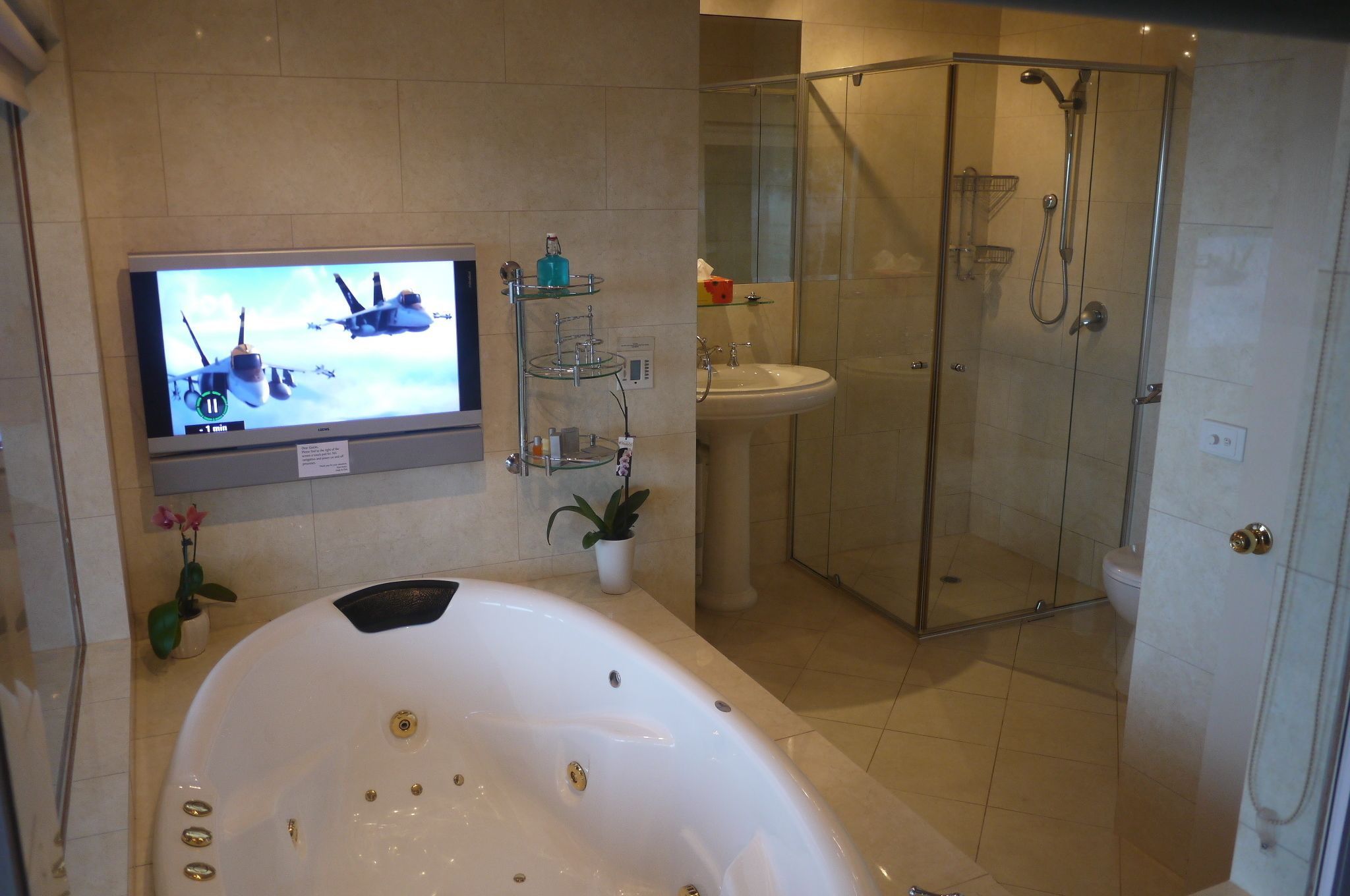 A modern bathroom with a glass-enclosed shower, a large bathtub, a sink, and a television mounted on the wall.
