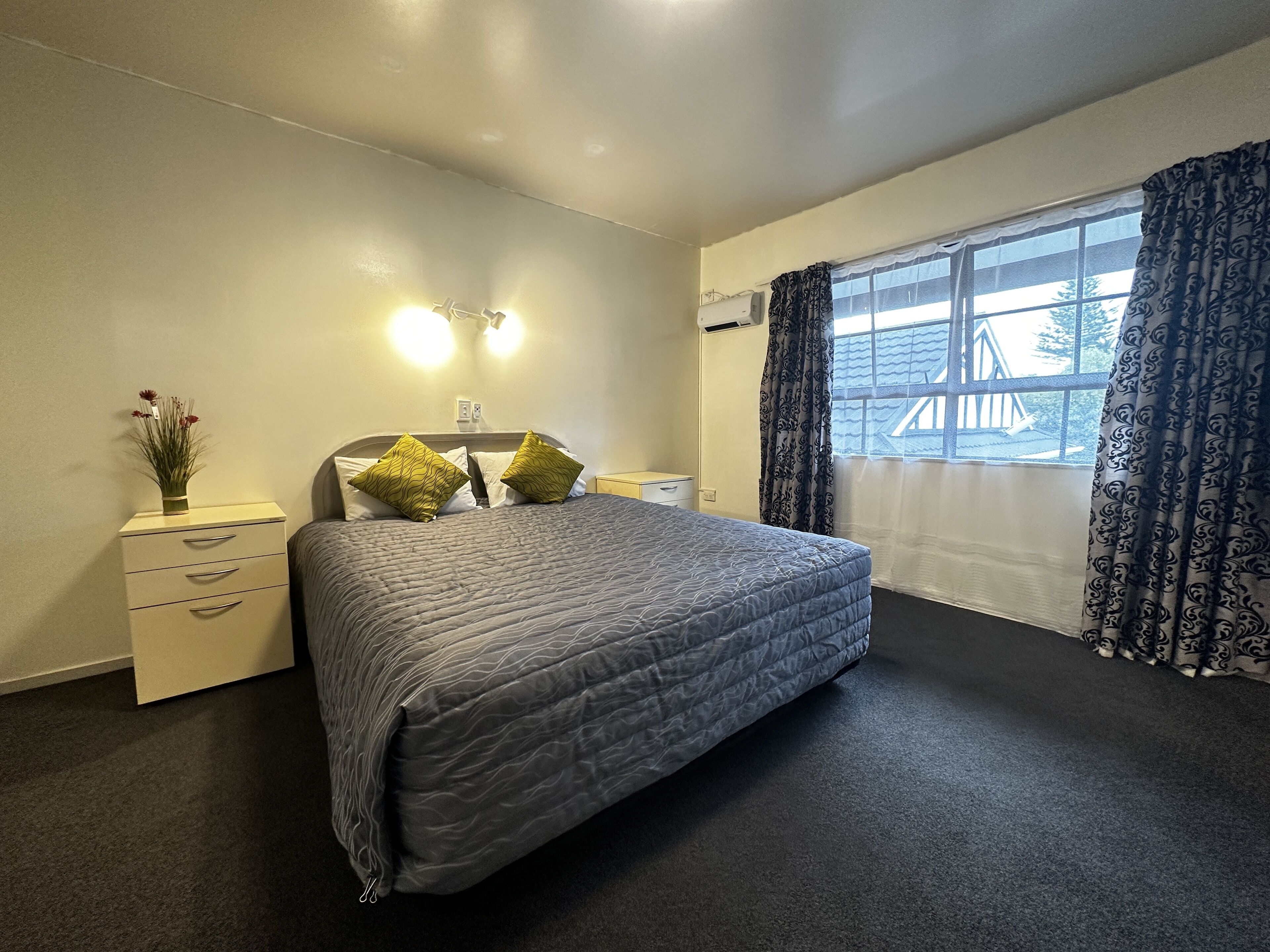 A bedroom with a bed, two bedside tables, a nightstand, a window with curtains, and a wall-mounted light fixture.