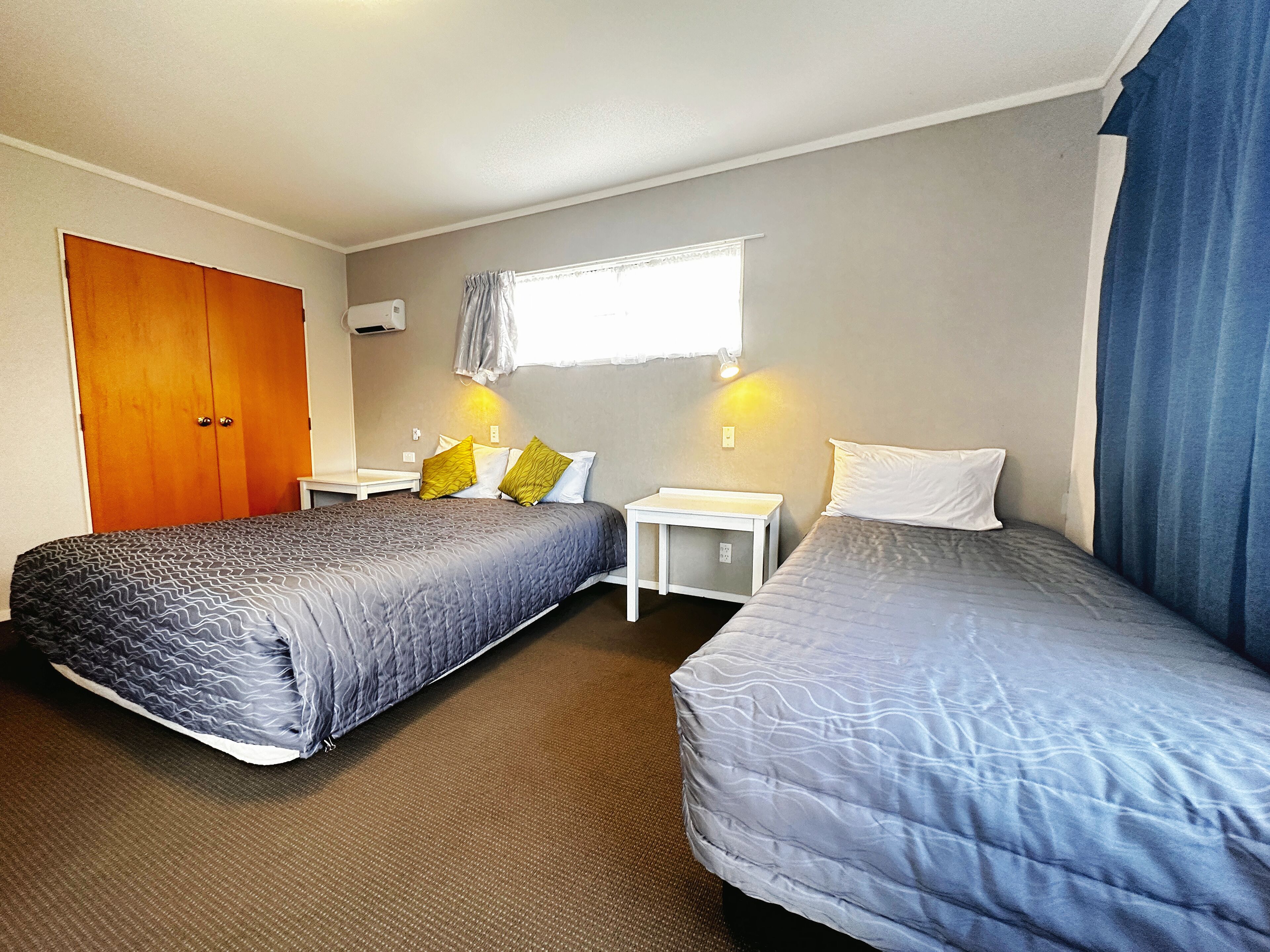 A hotel room with two beds, a wooden wardrobe, a small bedside table with a lamp, and a window with curtains.
