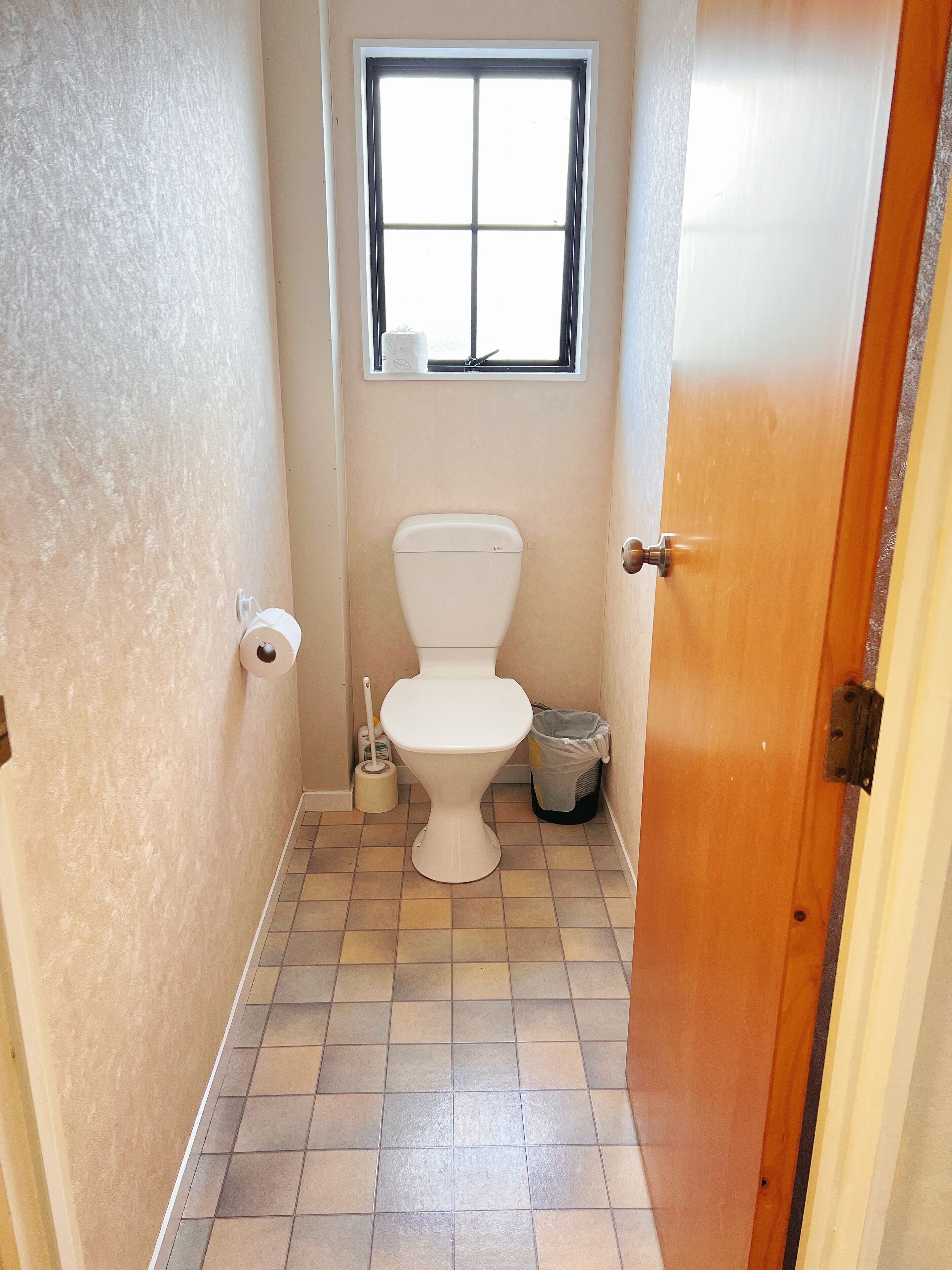 A small bathroom with a toilet, a window, a trash can, and a toilet brush.