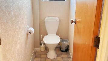 A small bathroom with a toilet, a window, a trash can, and a toilet brush.