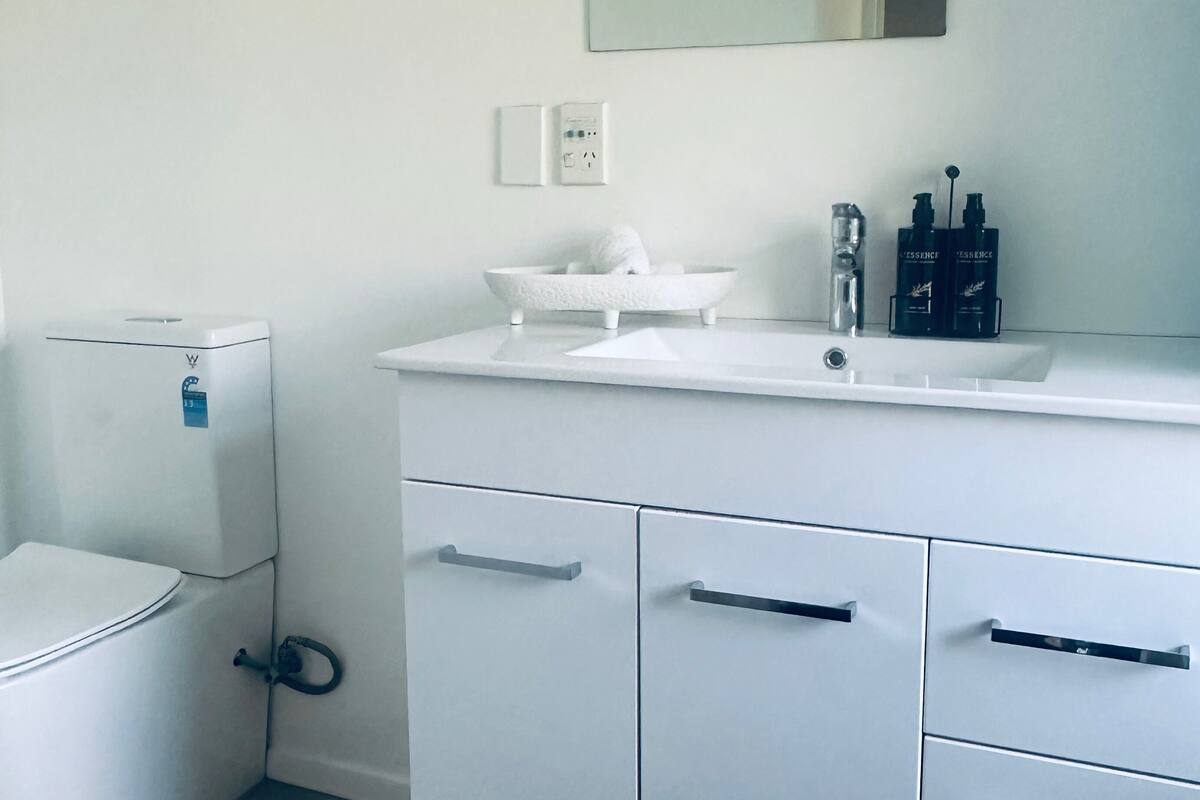 A modern bathroom with a white toilet, a vanity with a mirror, and a sink. There are toiletries on the sink and a towel hanging on the wall.