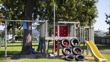 Children can play on the slide, climbing wall, and swing set in this shaded playground surrounded by trees and grass.