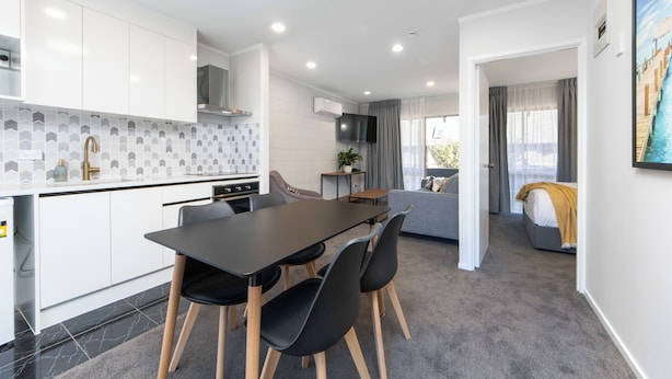 A modern kitchen with white cabinets, a black dining table, and a grey sofa. - Epsom Motel (Auckland)