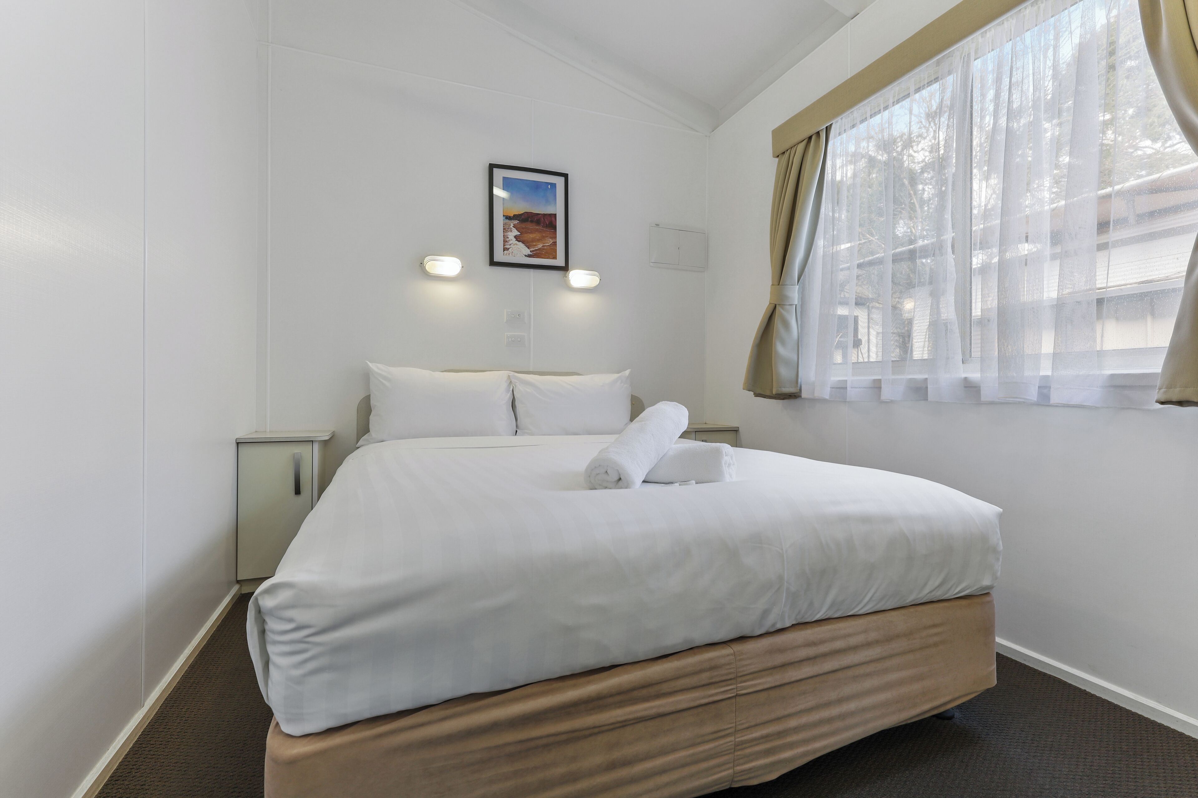 A hotel room with a bed, a nightstand, a wall-mounted picture, and a window with curtains.