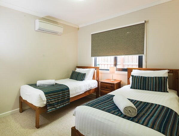A hotel room with two single beds, a wooden headboard, a nightstand, a lamp, and an air conditioning unit.