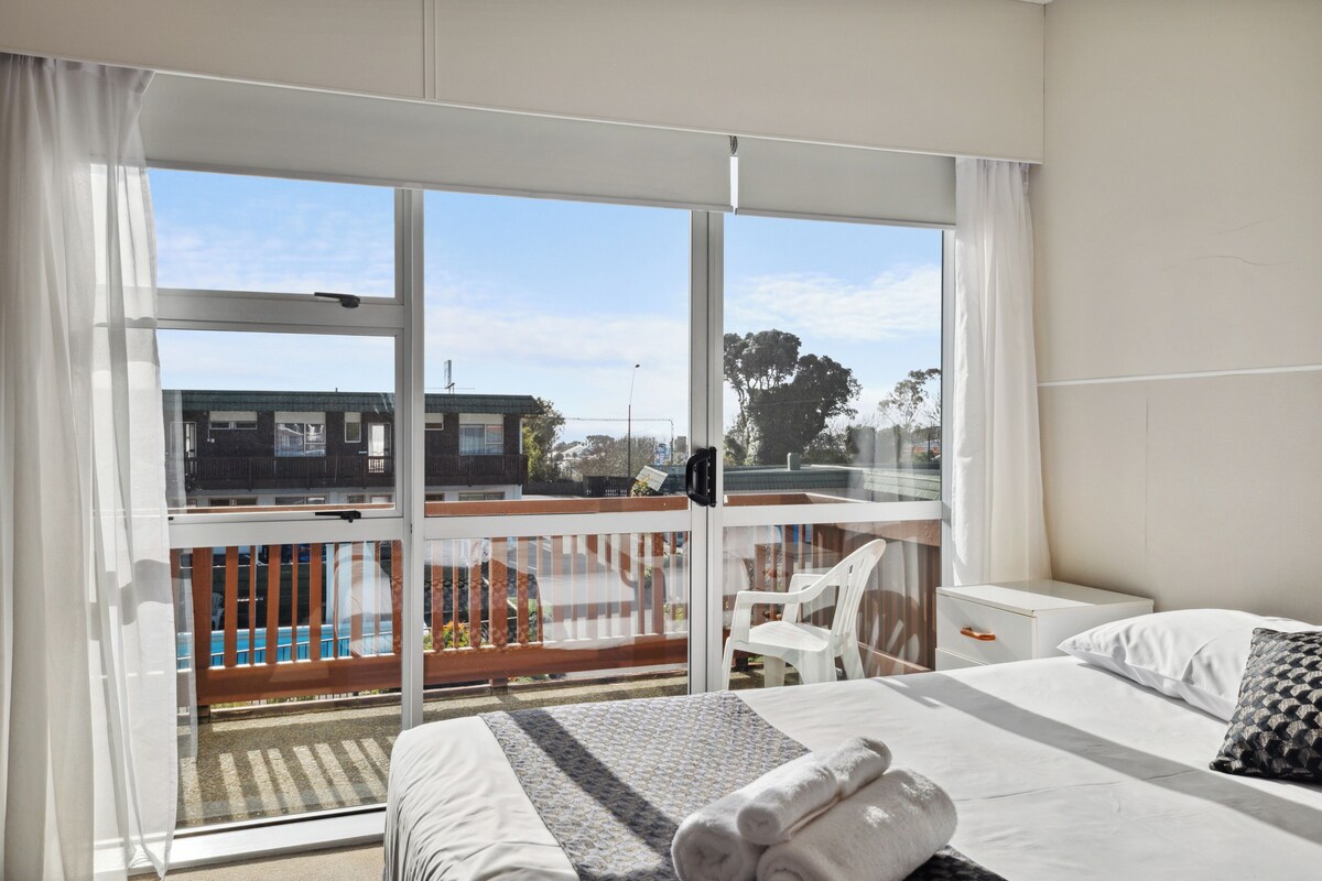 A hotel room with a large window, a bed with white linens, a small bedside table, and a view of a pool and buildings outside.
