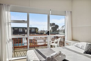 A hotel room with a large window, a bed with white linens, a small bedside table, and a view of a pool and buildings outside. - Flamingo Motel (New Plymouth)