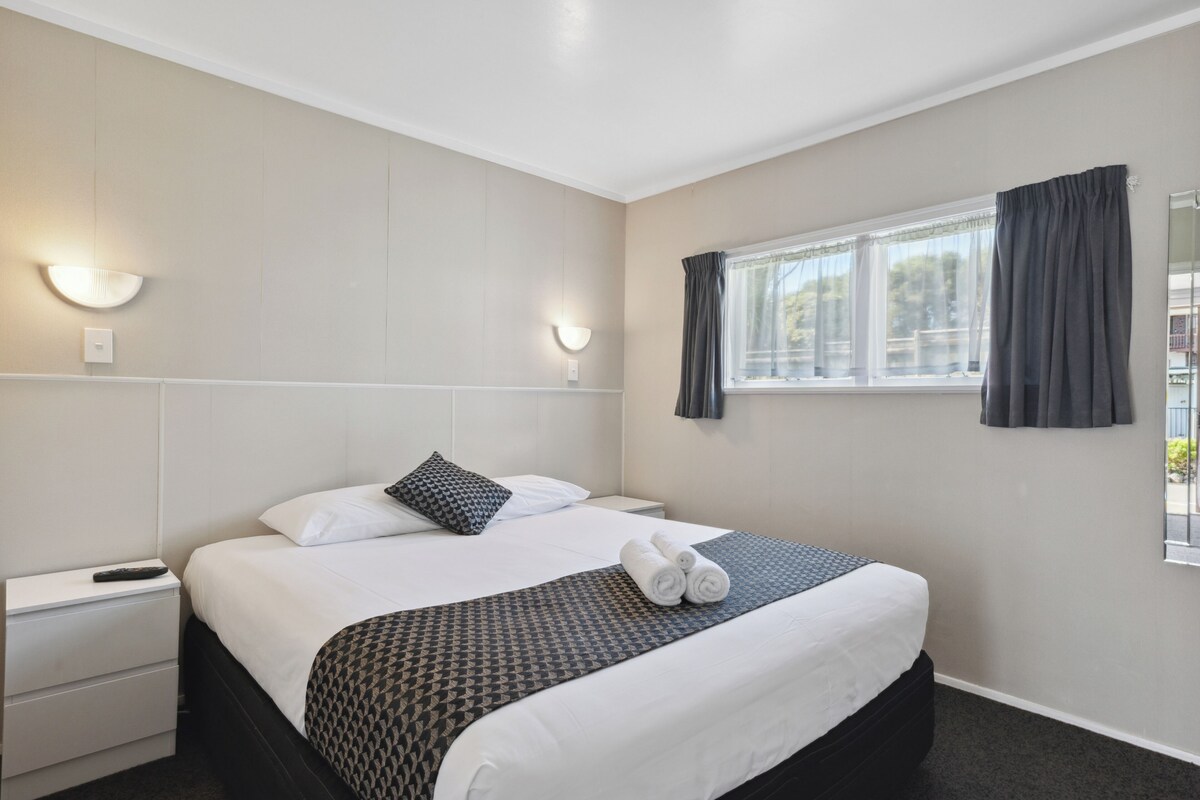 A hotel room with a neatly made bed, a bedside table with a remote control, and a window with curtains.