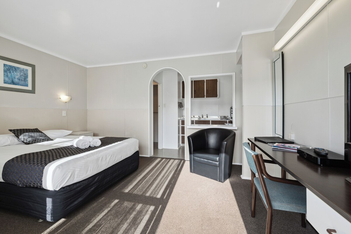 A hotel room with a bed, a desk with a chair, a mirror, and a small kitchenette.