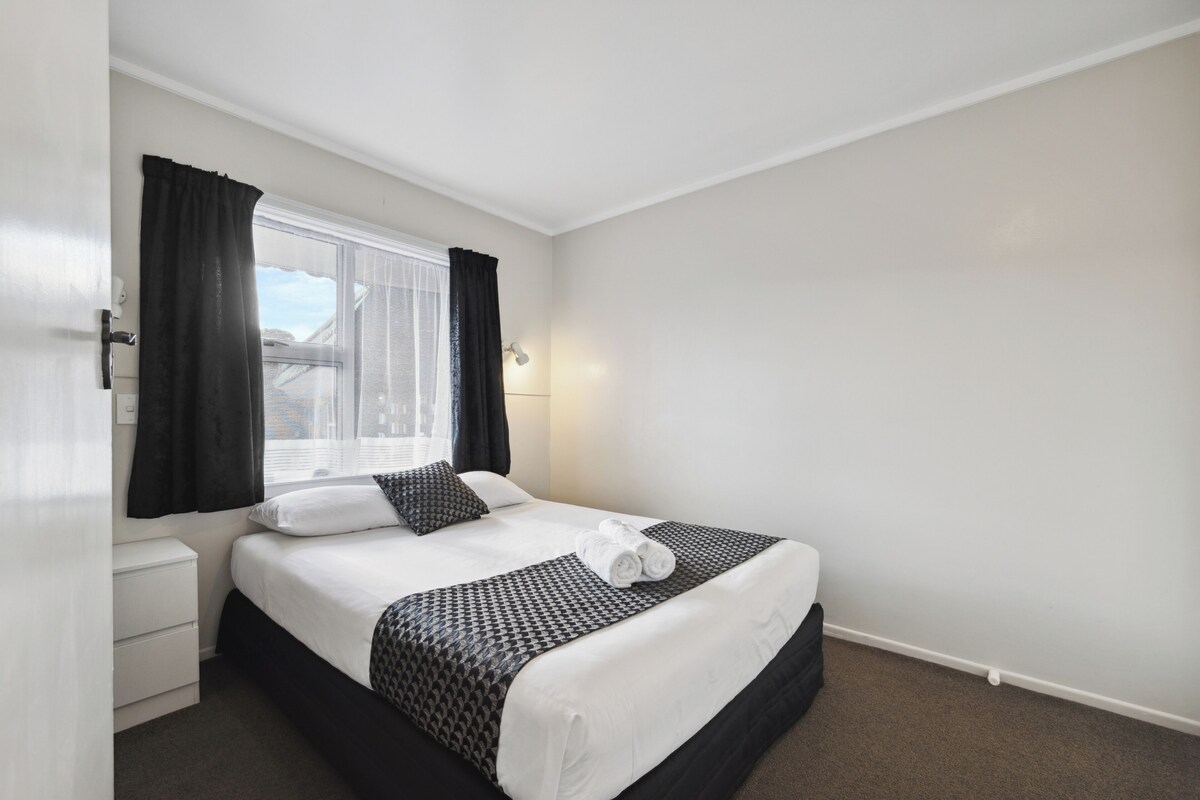 A neatly made bed with a patterned bedspread, a bedside table, and a window with curtains.