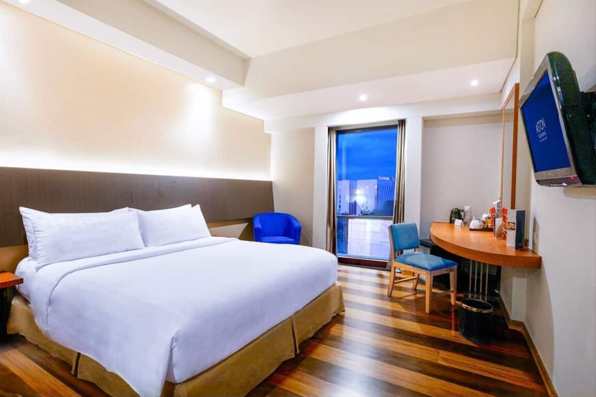 A hotel room with a large bed, a desk with a chair, a television, and a view of the city through a large window.