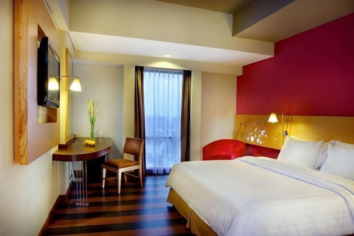 A hotel room with a bed, a desk with a chair, a TV, and a window with curtains.