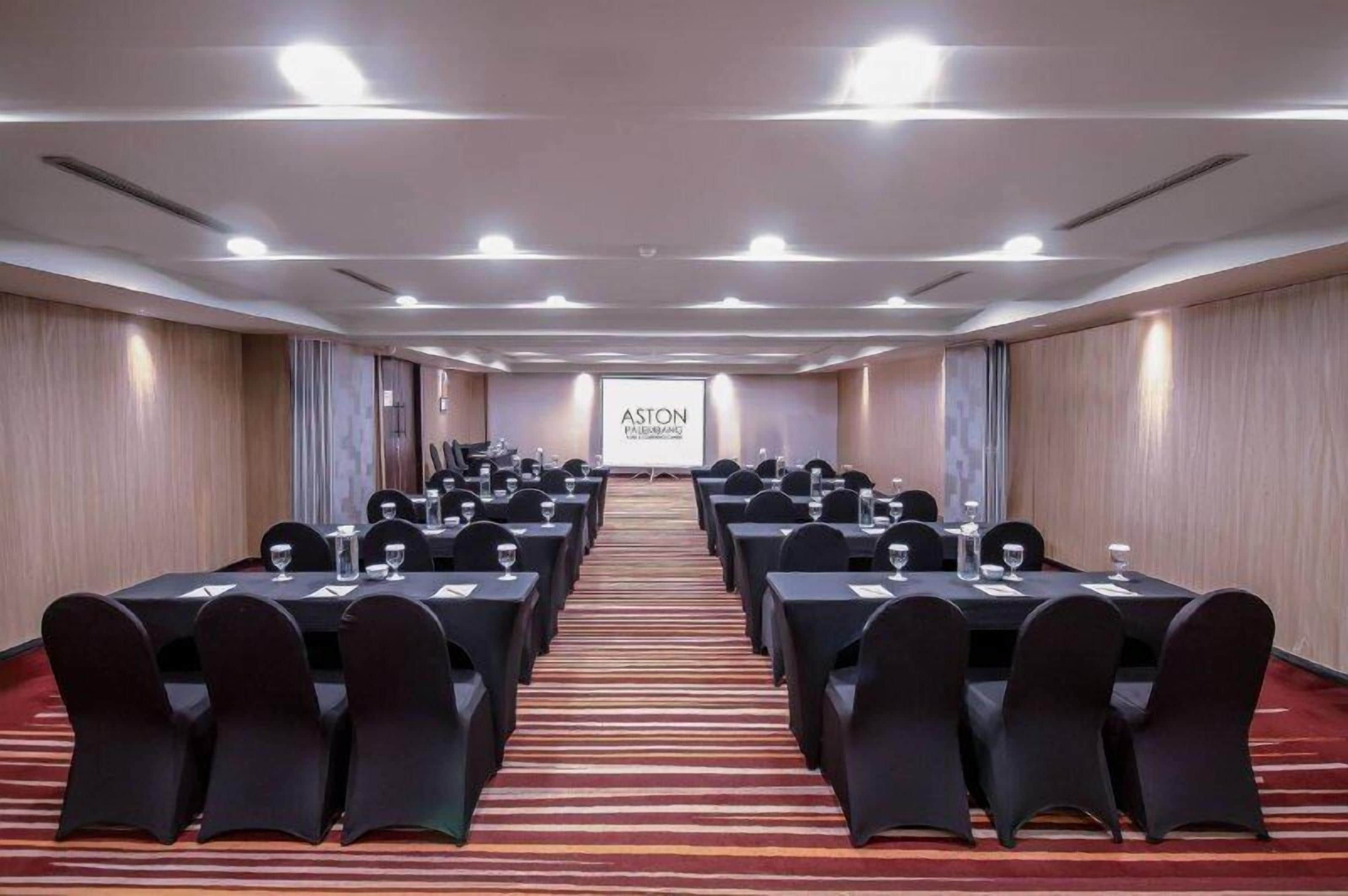 Un salón de conferencias con filas de mesas y sillas, piso alfombrado y una pantalla de proyección al fondo.