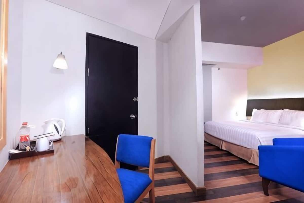 A hotel room with a wooden dining table, two blue chairs, a bed with white bedding, a black door, and a small white lamp.