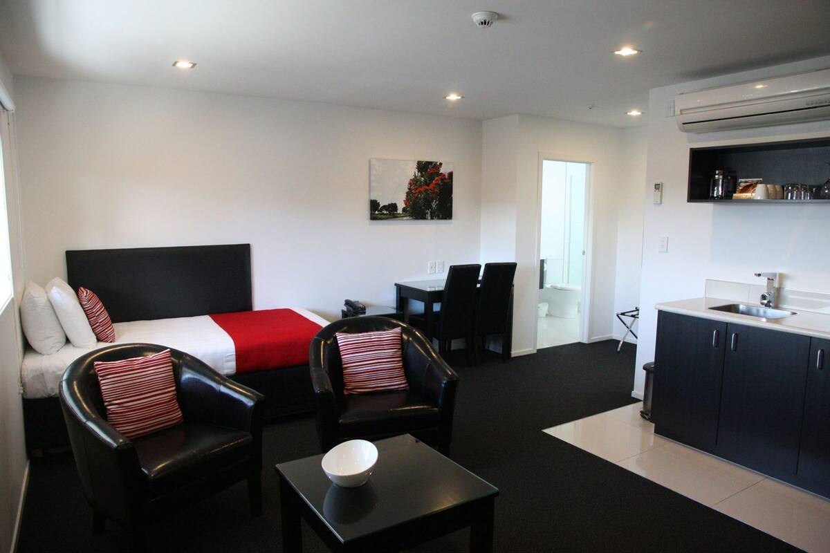 Habitación de hotel con cama, dos sillas, una mesita, zona de cocina y un cuadro en la pared.