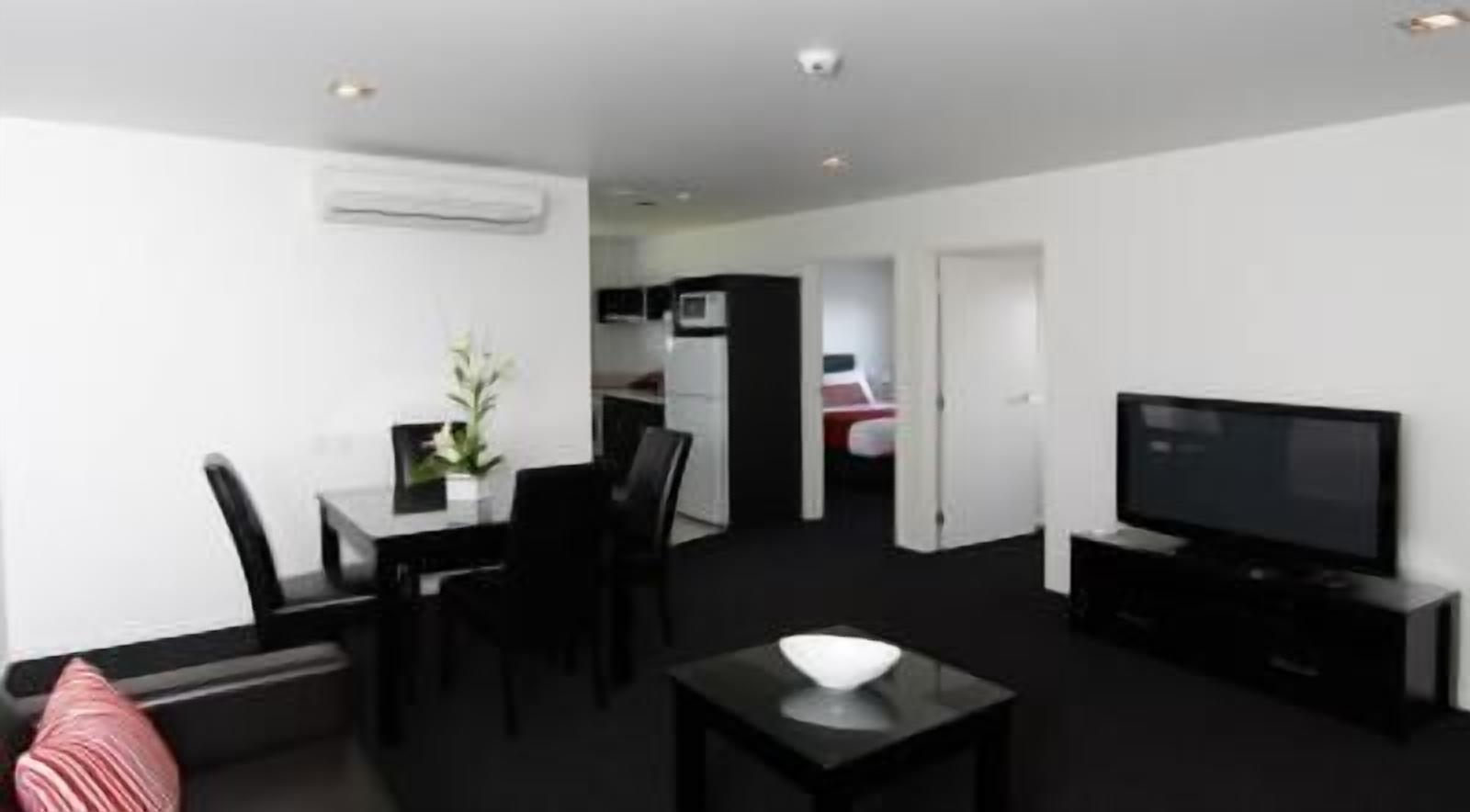 A modern living room with a flat-screen TV, a dining area with a table and chairs, and a kitchen in the background.