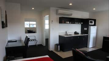 Quarto de hotel compacto com cozinha compacta, cama com detalhe em vermelho, uma cadeira e uma pequena lixeira.