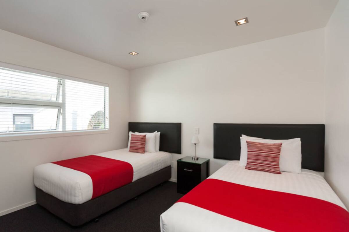 Habitación de hotel con dos camas, cada una con una colcha roja y blanca, una mesita de noche, una lámpara y una ventana con persianas.