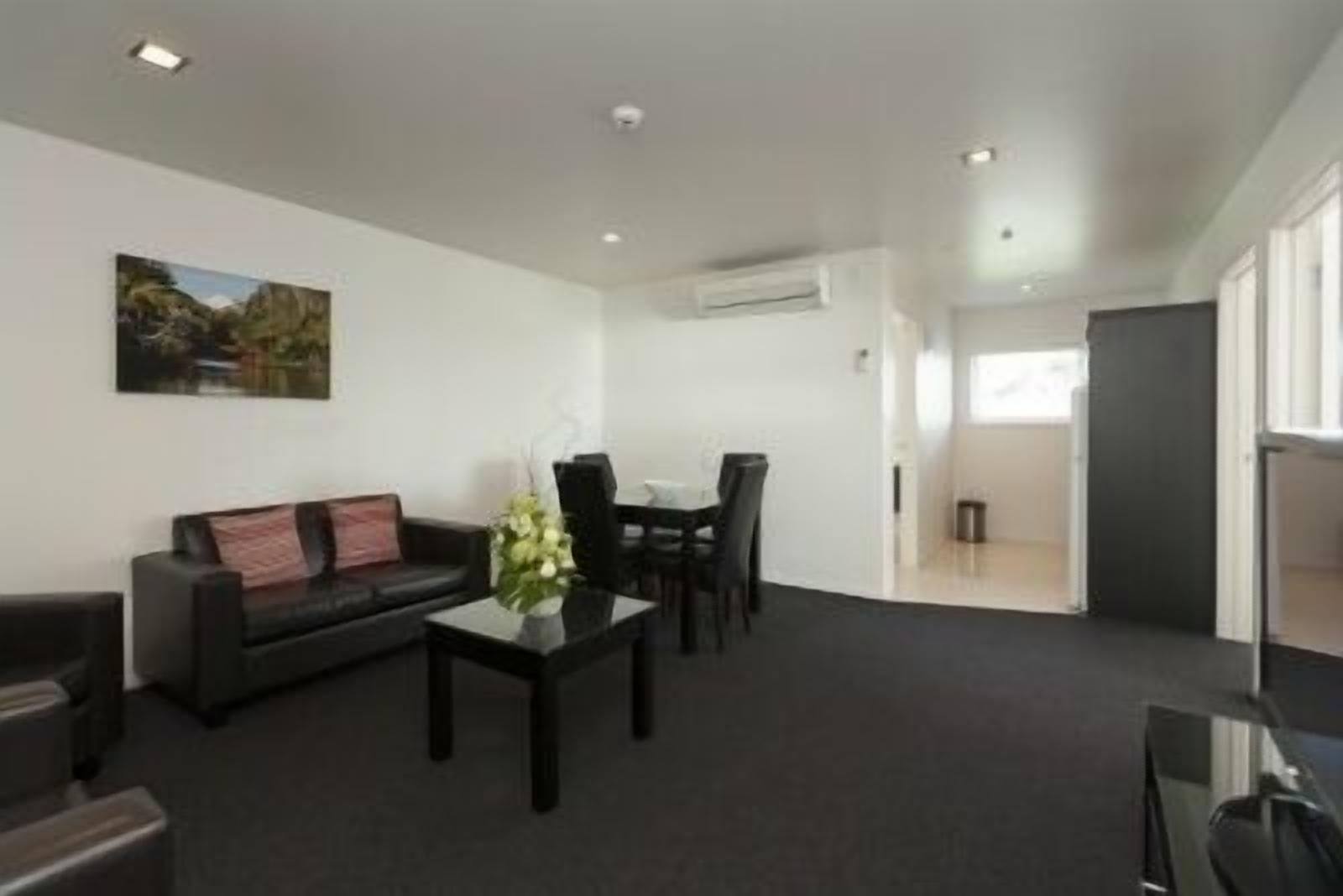 A modern living room with a black sofa, a coffee table, and a dining area with chairs. There is a painting on the wall and an air conditioning unit on the ceiling.
