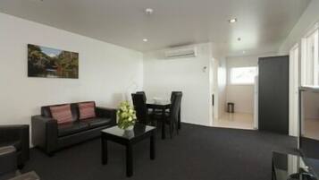 A modern living room with a black sofa, a coffee table, and a dining area with chairs. There is a painting on the wall and an air conditioning unit on the ceiling.