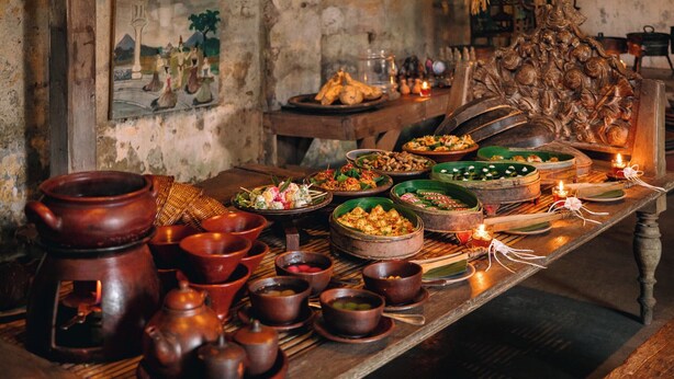 Een traditionele houten tafel met diverse gerechten en aardewerk, geplaatst in een rustieke kamer met stenen muren en een hanglamp.