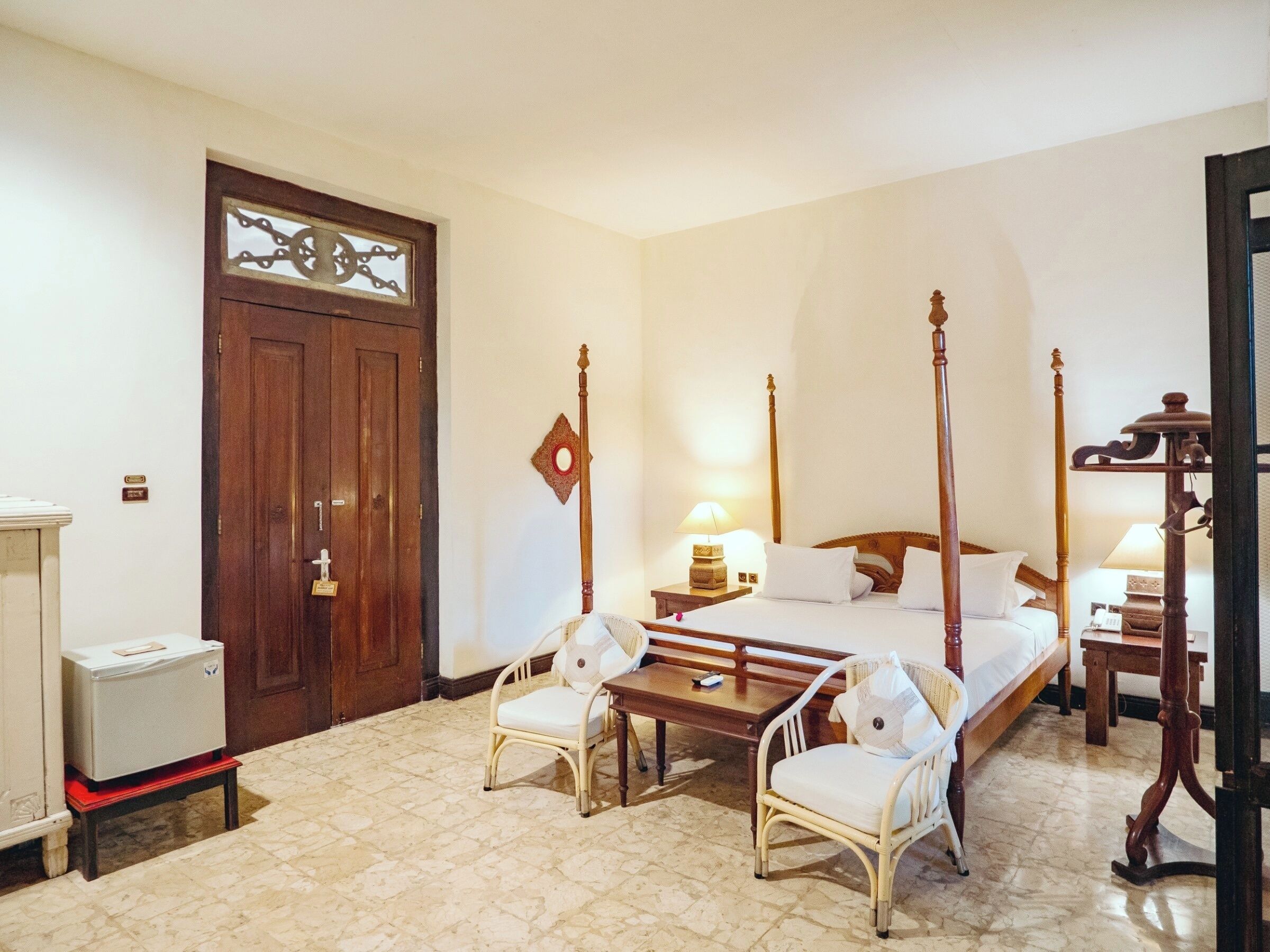Una habitación de hotel con una cama con dosel, dos sillas blancas, una mesa de madera, una mesita de noche con lámpara y un refrigerador.
