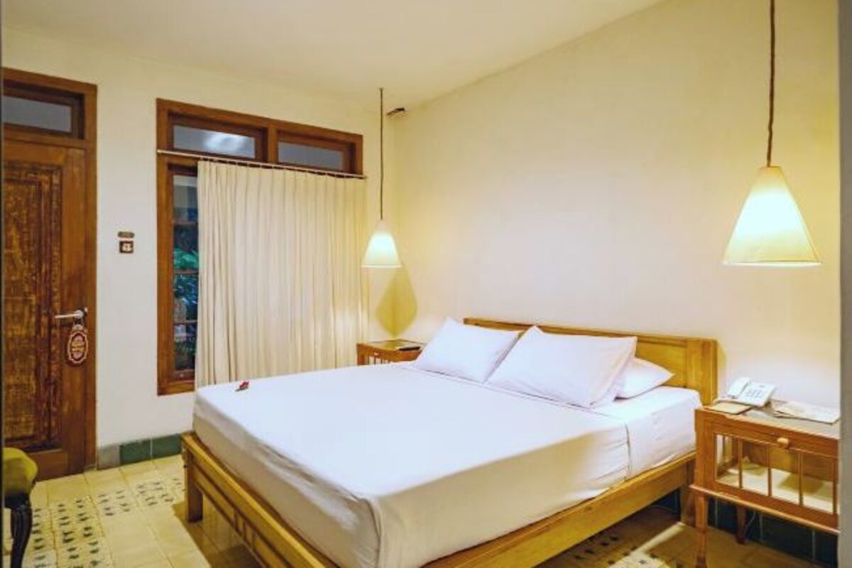 Un dormitorio con cama de madera, ropa de cama blanca, una mesita de noche con lámpara y una ventana con cortinas.