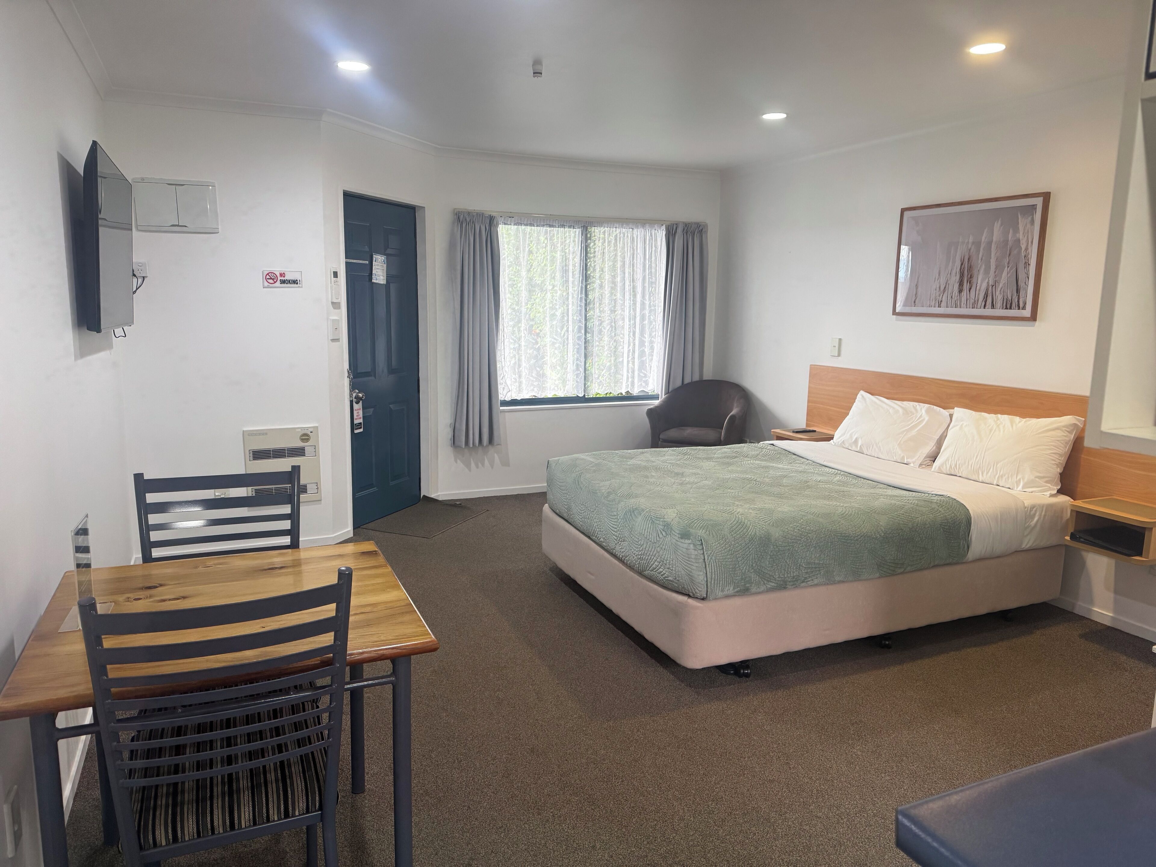 Habitación de hotel con cama, escritorio con silla, ventana con cortinas, televisor montado en la pared y un cuadro enmarcado.